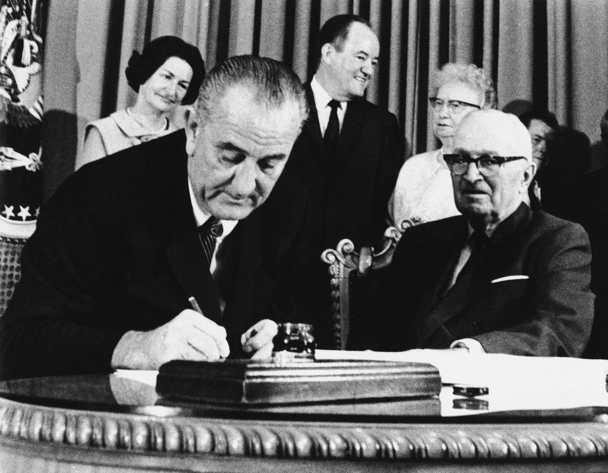 President Lyndon Johnson signs Medicare into law on July 30, 1965, while ex-President Harry S. Truman, right, observes at the Truman Library in Independence, Mo. At rear are Lady Bird Johnson, Vice President Hubert Humphrey and former first lady Bess Truman. (Associated Press)