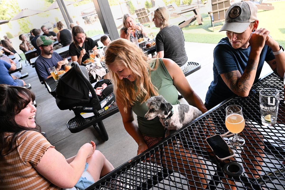 Briana Stroh, left, a beertender at Uprise Brewing Co. in Spokane, laughs as she offers Milo a dog a treat while chatting with Milo’s dog parents CK Daily, center, and Bob Maureen on July 14 at Uprise  (Tyler Tjomsland/The Spokesman-Review)