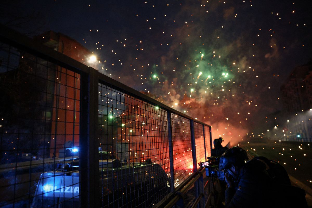 A person uses a camera while standing behind a fence next to a police vehicle, with fireworks exploding as demonstrators use them against police officers while trying to block a road leading to the Santa Giulia Arena, on the day of a protest against the environmental, economic and social impact of the Milano-Cortina 2026 Winter Olympics, in Milan, Italy, February 7, 2026. REUTERS/Guglielmo Mangiapane (Guglielmo Mangiapane)
