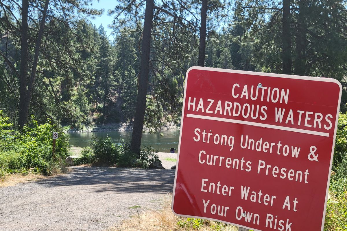 Large red signs near the shore of the Spokane River in Corbin Park advise visitors to take caution if entering the water. The park is a popular spot for water recreation.   (Nick Gibson / The Spokesman-Review)