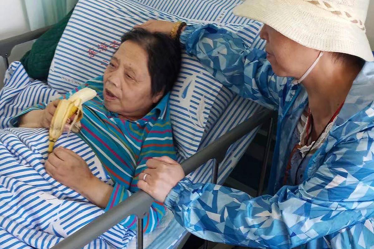 In this photo released by the family of Shen Peiming, Shen Peiming, 71, eats a banana as a family member attends to her at her bed side at the Shanghai Donghai Elderly Care hospital on Sept. 24, 2019. Shen died Sunday morning, April 3, 2022, at the hospital, without her loved ones by her side. Her family, unable to visit because of pandemic restrictions, is unsure of the circumstances of her death. The hospital had reported a COVID-19 outbreak, but Shen had tested negative, as of last week.  (HONS)