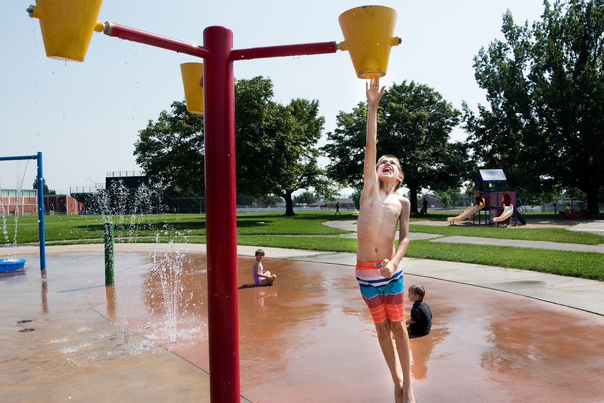 Zachary Jordan leaps to tip a bucket of water at the Shadle Park Splash Pad on Aug. 8, 2018, as his sister Emmaline and little brother Henry play in the water.  (TYLER TJOMSLAND/THE SPOKESMAN-REVIEW)