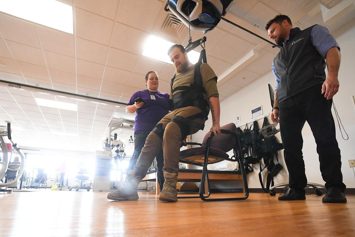 Neurophysical therapist Sarah Gross, left and Jake Allstot, right, Assistant manager of rehabilitation at St. Luke's help Cameron Tweedy, 21, center, out of a chair with the aid of a ZeroG Gait and Balance System during a staged demonstration of the machine and Tweedy's recovery process on Thursday, Jan. 30, 2020, at St. Luke's Rehabilitation Center in Spokane, Wash. According to the manufacturer's website, the "ZeroG Gait and Balance System by Aretech uses dynamic body-weight support and fall protection for intensive gait and balance therapy." (Tyler Tjomsland / The Spokesman-Review)