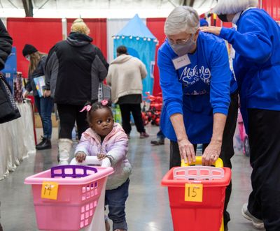 Volunteer Sandy Richardson, right, walks with Eva Akon, 23 months old, with a shopping cart while her parents shop for Christmas gifts in the toy room at the Christmas Bureau on Tuesday, Dec. 7, 2021, at the Spokane Fair and Expo Center in Spokane. Eva’s family is originally from South Sudan. After operating remotely in 2020 because of COVID-19, the annual charity that gives away toys and food vouchers is back again with health protocols in place. The annual event offers a high-quality toy for each child and a gift certificate for groceries to every family, regardless of income.  (Jesse Tinsley/THE SPOKESMAN-REVIEW)